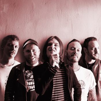 Preview: Bansfoto Bakemono Stereo: Fünf Personen stehen eng nebeneinander vor einer hellen Wand. Das Foto ist in warmen Rot- und Sepiatönen gehalten. Die Personen tragen überwiegend dunkle Kleidung, einige haben kurze Haare, eine Person hat schulterlanges Haar. Di